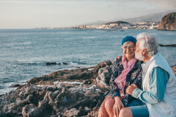Romantic smiling senior couple sitting on the rocky beach at sea enjoying vacation and retirement...