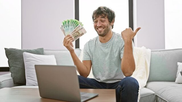 Cheerful young man at home, confidently flashing a thumbs up while holding russian rubles, with a joyous open-mouthed smile, pointing to the side with his laptop.