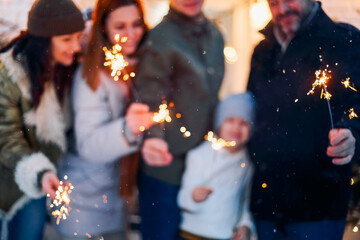 Portrait of big happy family with child standing in snow-covered house yard holding lit sparklers in hands. Parents and children of different age rejoicing Christmas and New Year holidays outdoors