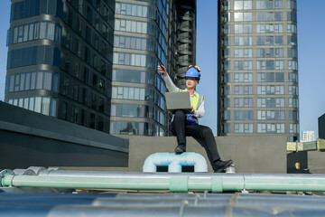 Engineer on building rooftop with laptop, overseeing urban construction, blue sky, and skyscrapers.