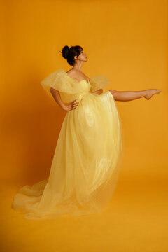 A woman in a yellow dress is posing for a photo. The dress is long and has a polka dot pattern. The woman is standing on one leg, and the photo is taken on a yellow background.
