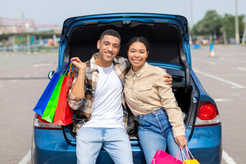Positive young couple in casual wear posing near their car and smiling at camera, holding shopping bags, feeling happy