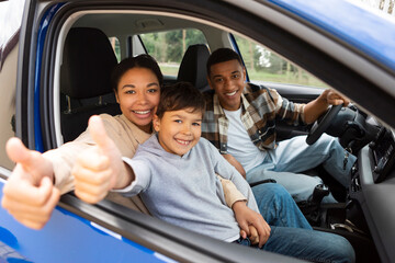 Happy family of three, mother, son and father driving car, woman and boy showing thumbs up in open window, enjoying time together at weekend