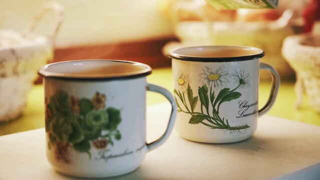 Tea time with enamel cups. Two rustic enamel cups, featuring flower designs, are placed on the table. Countryside house. Tea with milk. 4K