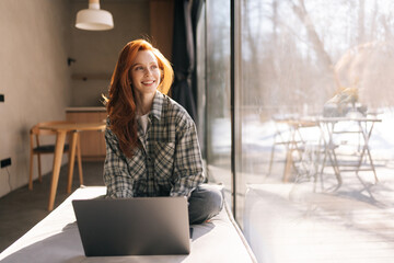 Portrait of happy freelancer female remote working on laptop sitting on wide windowsill with nature view on sunny day. Cheerful young student typing on laptop keyboard, using online app at home. - obrazy, fototapety, plakaty