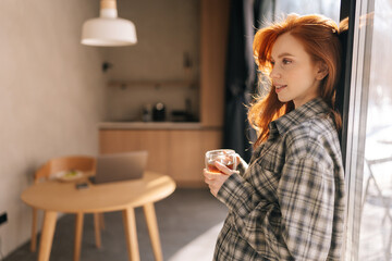 Side view portrait of pretty young woman holding cup enjoy smell coffee or tea and drinking with happiness standing by window on sunny morning. Smiling female enjoys good morning feels stress free.