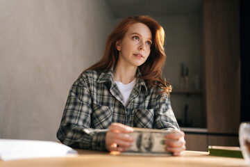 Low-angle view of thoughtful young woman holding banknotes feeling stress about unemployment at home workplace. Upset female holding money and disappointed about bad earnings salary.