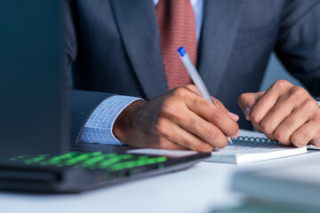 Business man wearing blue color suit and red tie writing on notepad, working on laptop
