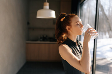 Side view of athletic young woman in sport outfit standing by window and drinking fresh water in morning after fitness training workout exercise at home, enjoying sunny weather and lifestyle.