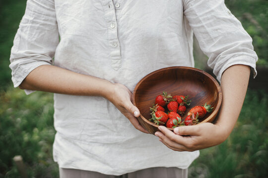 Woman holding wooden bowl with strawberries close up. Gathering fresh natural berries in urban organic garden. Homestead lifestyle