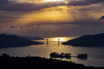 A suspension bridge during sunrise or sunset, with golden hues reflecting off the calm water. Small islands and hills are in the background.