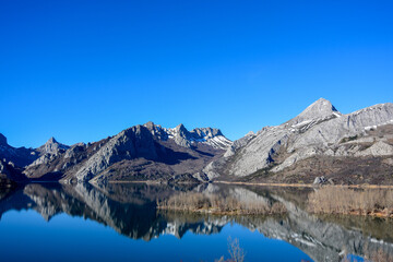 Rugged mountains with sharp peaks and ridges reflecting perfectly in a calm, clear body of water. The vibrant blue sky above enhances the natural beauty and serene atmosphere of the landscape.