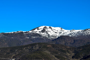 A majestic snow-capped peak under a clear blue sky, with lower, darker hills in the foreground. Sparse vegetation contrasts the white snow and blue sky, highlighting the natural beauty of the area.