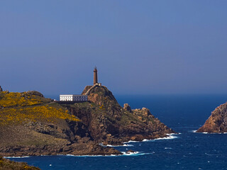 A tall cylindrical lighthouse stands on a rocky promontory, with a white keeper's house below. Waves crash against the rocks under a clear blue sky, highlighting the rugged coastal landscape.