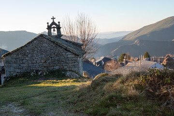 A small stone chapel with a cross on top, set in a rural, mountainous area. Surrounded by grass and bare trees, with a village and rolling hills in the background, bathed in soft light.