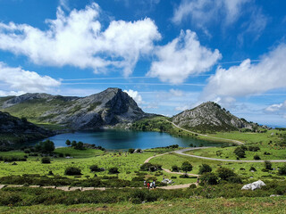 A serene lake surrounded by lush green fields, rolling hills, and rocky mountains under a blue sky with clouds. A winding road leads to the lake, with people in the foreground enjoying the scenery.