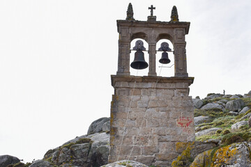 A stone bell tower with two bells in separate arches, situated on rocky terrain with moss and lichen. The tower has a cross at the top and red markings on the lower right side. The sky is overcast.
