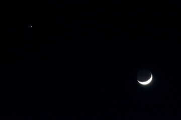 Crescent moon seen in a night sky with a single star in the distance
