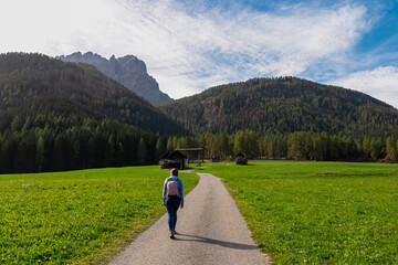 Woman on scenic hiking trail along wooden huts on idyllic alpine meadow with view of majestic mountain peaks of Sexten Dolomites, South Tyrol, Italy, Europe. Hike panoramic Fischleintal, Italian Alps - obrazy, fototapety, plakaty