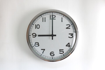 A round shape stainless steel wall clock with white face and hands set to 9 o'clock time. The clock is hanging on a wall.