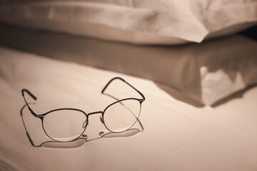 A pair of glasses is laying on a bed. The bed is unmade and the pillows are stacked on top of each...
