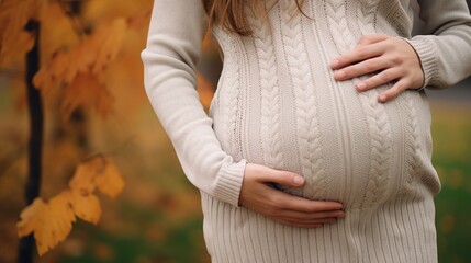 Close up of pregnant woman in sweater touching her belly in the autumn park