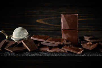 Broken pieces of dark chocolate bar on a black sorting board and a kitchen strainer for chocolate chips. Advertising space