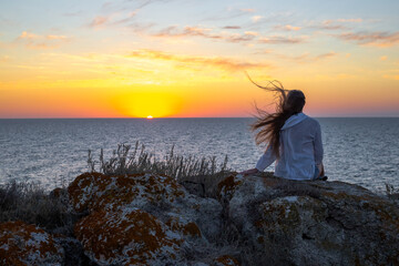 A woman with long hair sits on a stone on the top of a mountain on the sea coast at sunset, rear view. Travel and tourism.