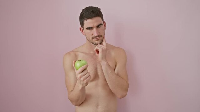 Clueless young hispanic man, shirtless with apple in arms, displays bewildered 'no idea' expression over isolated pink background.