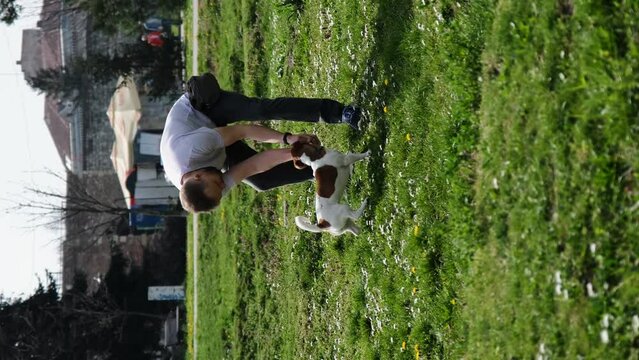 Caucasian man plays with dog in green clearing. Male pet owner with a Jack Russell terrier on a walk on a sunny spring day, actively and having fun. Vertical 4k footage with happy pet