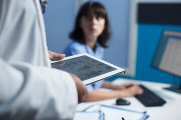 Selective focus on an individual wearing a white lab coat grasping a tablet while a nurse uses a...