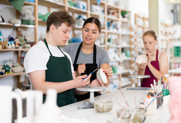 Young woman teacher showing teenage boy and teenage girl how to paint ceramic cup in workshop