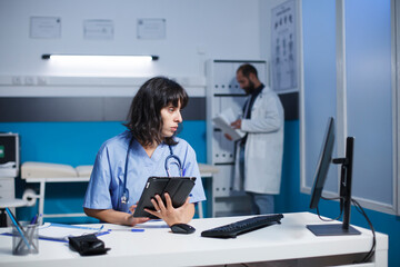 Obraz premium Female nurse uses a smart tablet and computer for medical information and checkups. While examining patient data on desktop pc, healthcare professional holds a gadget with a touch screen.