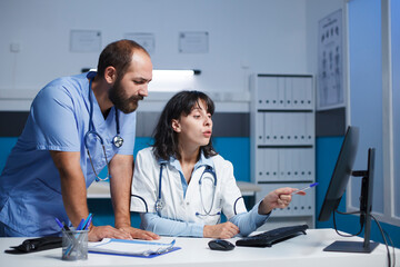 Caucasian medical staff in blue scrubs and lab coat collaborate digitally in a clinic office. They...