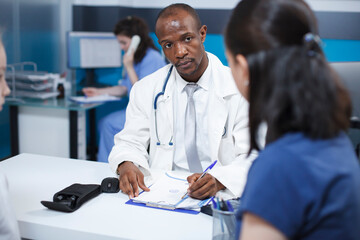 Black man wearing a lab coat, taking notes during a consultation with a female patient at the...