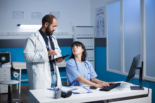 Caucasian doctor and female healthcare worker collaborate in a clinic office. They discuss digital medical images on a tablet and desktop computer. Medical equipment is visible.