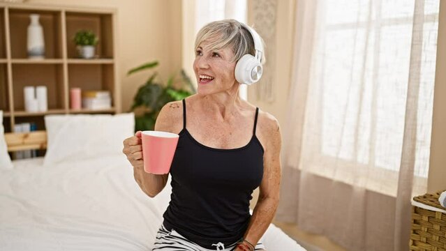 A mature woman with grey hair listens to music in a cozy bedroom while holding a pink mug, embodying relaxation and contentment.