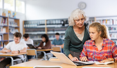 Friendly female teacher helps schoolgirl find information in laptop in the school library