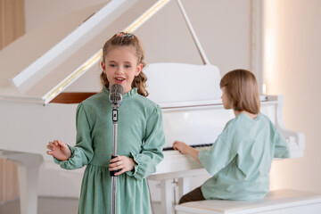 girl sings into a microphone, a boy accompanies her on the piano.