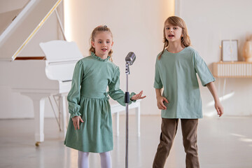Two children sing into a microphone against the backdrop of a white piano.