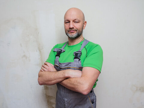Portrait of a construction worker on light wall background. Builder in grey heavy duty clothes and green shirt. Hands crossed. Male in 40s, bald with grey beard. Lean athletic type.