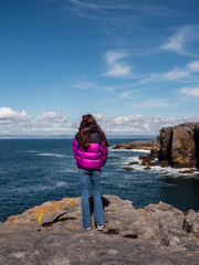 Teenager girl enjoys stunning view of mini cliffs in county Clare, Ireland. Warm sunny day. Travel and tourism. Irish landscape. Model with long hair, red jacket, slim body type.