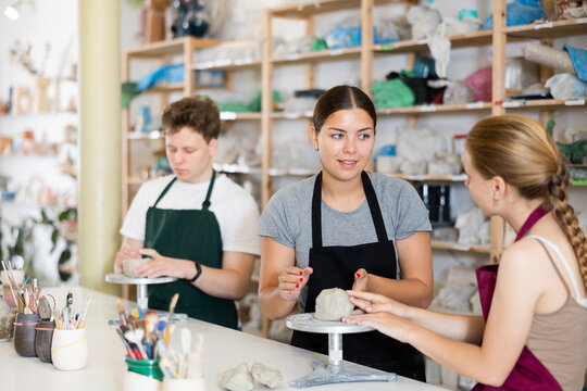 Young female teacher in apron helps teenager boy and teenager girl students to make product from clay in ceramic workshop