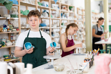 Young man in an apron demonstrates clay mugs, which he himself painted with colored paints