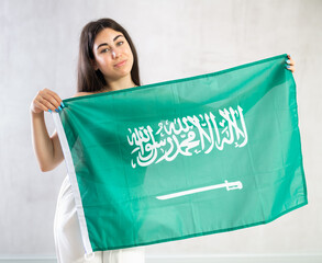 Cheerful young woman standing with big flag of Saudi Arabia against light shadeless background