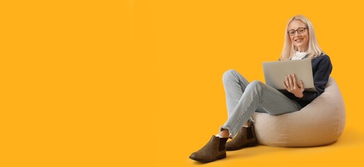 Mature female programmer with laptop on beanbag chair against yellow background with space for text