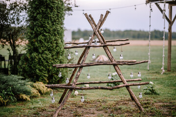 Valmiera, Latvia- July 28, 2024 - A rustic wooden shelf in an 'X' shape with hanging glass vases, set against an outdoor backdrop with greenery and a string of lights.