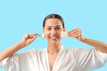 Beautiful young woman with facial massage tools on blue background, closeup