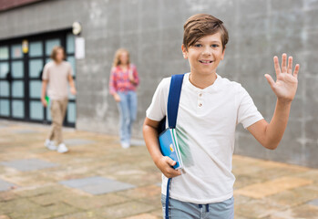 Portrait of positive school boy standing in front of a school building and waving hand somebody