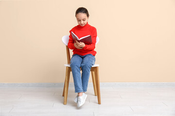 Little girl reading book while sitting on chair near beige wall